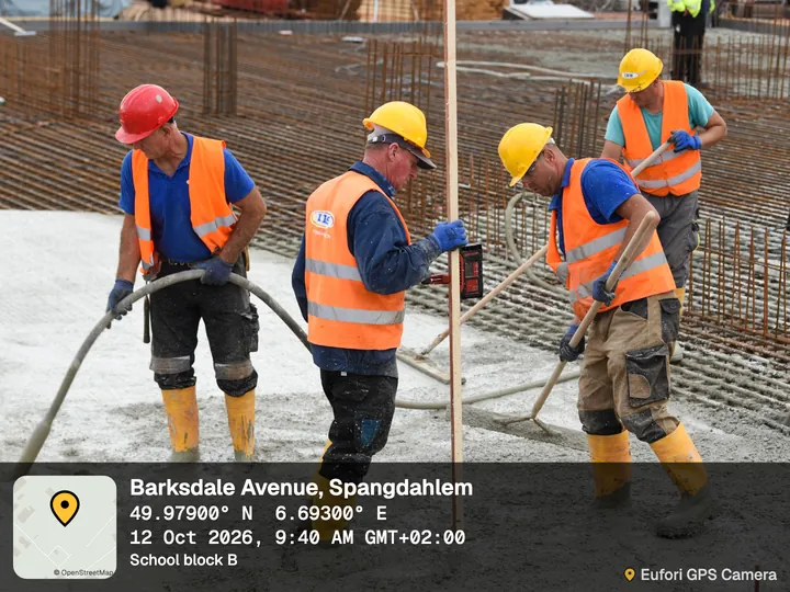 Crew in hard hats pouring concrete over rebar, with the app's Classic stamp: Barksdale Avenue, Spangdahlem, coordinates, date and time, project School block B